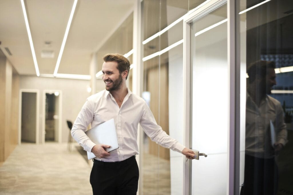 pexels-photo-901424-901424 A smiling man holds a laptop while standing in a modern office hallway, exuding confidence.